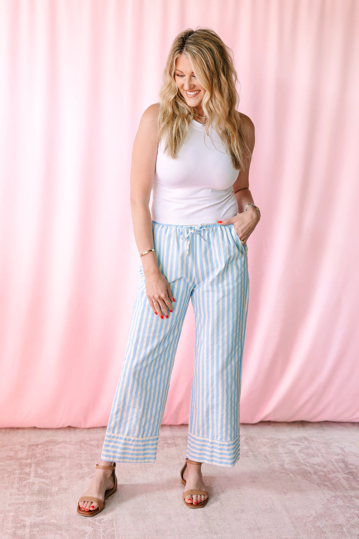 Woman smiling wearing blue and white striped wide-leg crop pants with a white sleeveless top and tan block heel sandals against a pink backdrop
