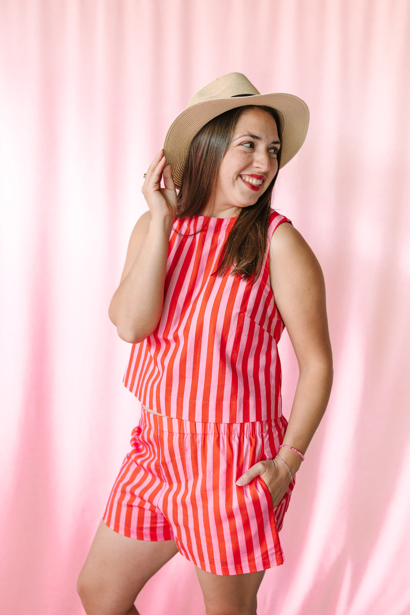 Smiling woman wearing a pink and coral vertical stripe sleeveless top with matching striped shorts, styled with a tan wide-brim hat against a pink backdrop