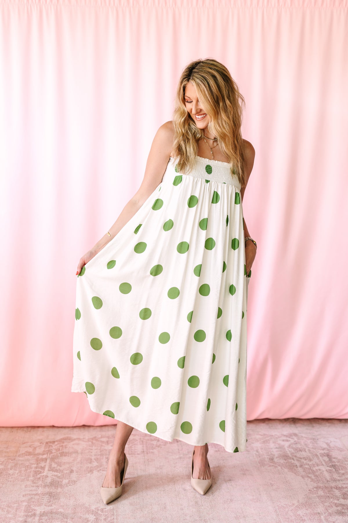 Full-length view of smiling woman in a white and green polka dot smocked maxi dress with flowing A-line skirt and nude pointed-toe heels against a pink backdrop