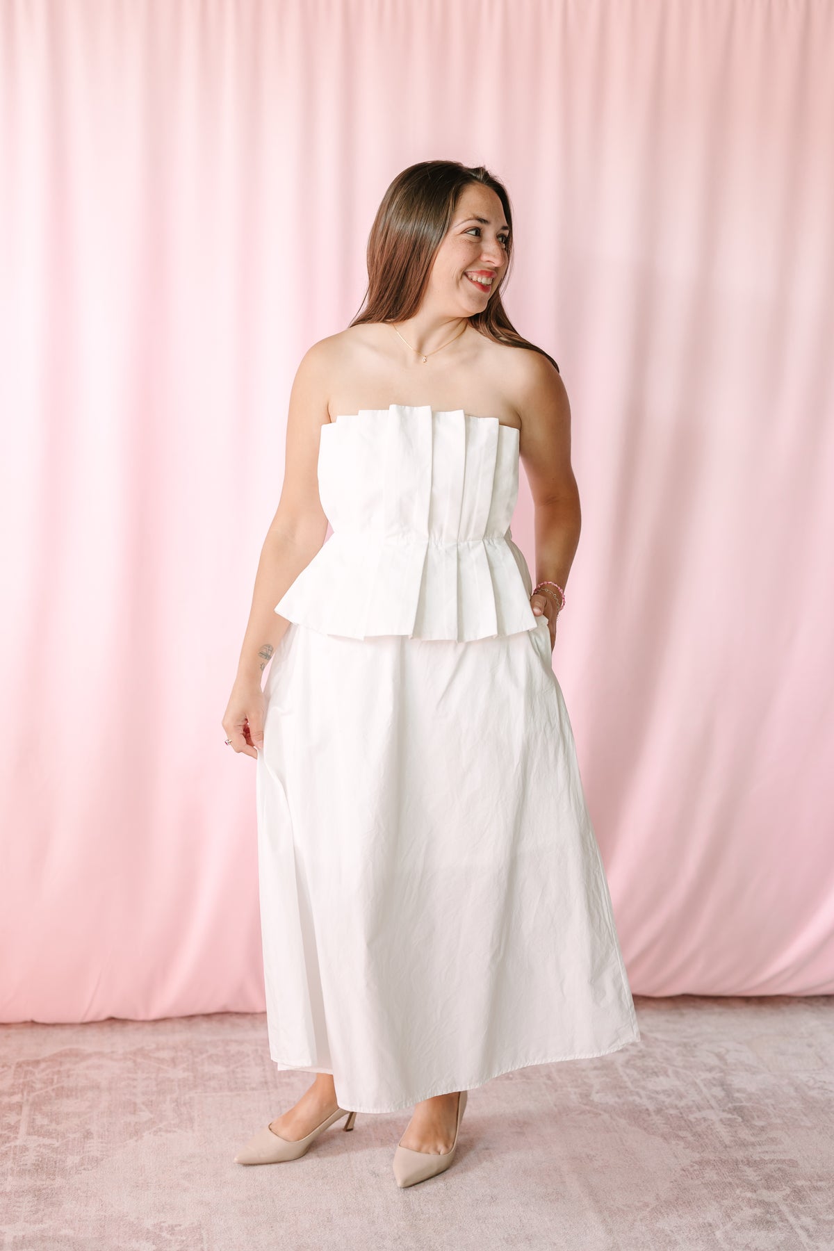 Full-length view of woman in white strapless pleated peplum dress with nude pointed-toe heels against a pink backdrop