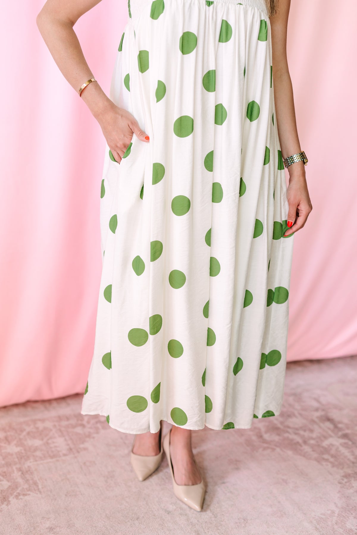 Close-up of white and green polka dot flowing maxi skirt with side pocket detail and nude pointed-toe heels against a pink backdrop
