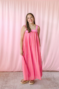 Smiling woman wearing a pink multicolor vertical stripe sleeveless maxi dress with square neckline and wide straps, styled with tan platform sandals against a pink backdrop