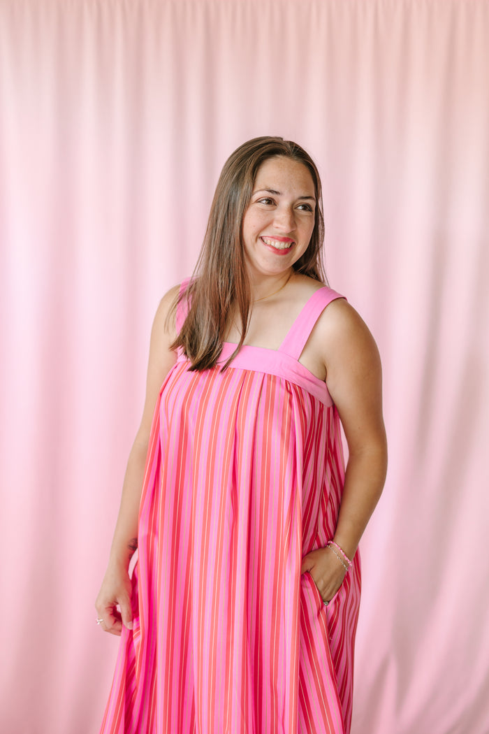 Woman smiling in a pink multicolor vertical stripe sleeveless maxi dress with hands in pockets, showing relaxed A-line silhouette against a pink backdrop