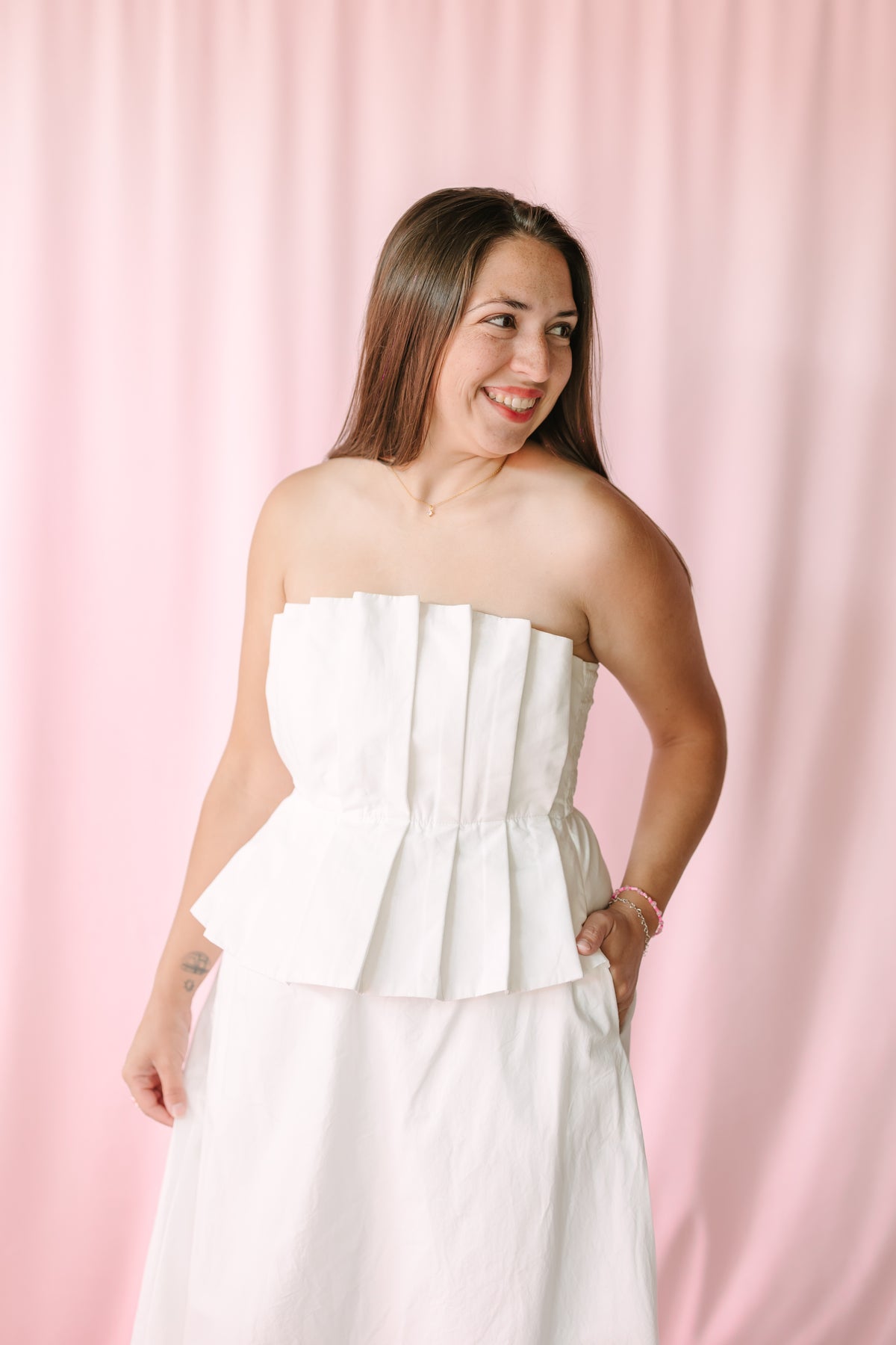 Smiling woman wearing a white strapless pleated peplum dress styled with gold jewelry against a pink backdrop