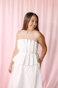 Smiling woman wearing a white strapless pleated peplum dress styled with gold jewelry against a pink backdrop