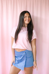 Smiling woman in a blue and white gingham skort and pink crop tee, three-quarter view against a pink backdrop