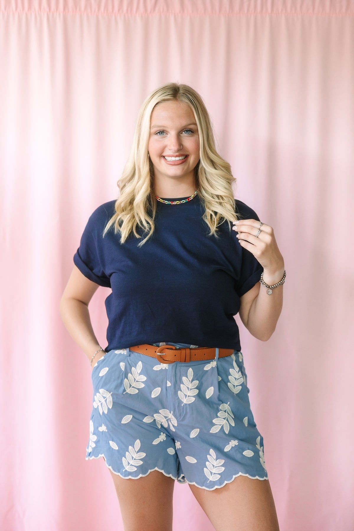 Woman wearing blue embroidered leaf shorts with scalloped hem, navy t-shirt, and tan leather belt, styled with nude heeled sandals
