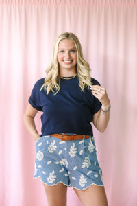 Woman wearing blue embroidered leaf shorts with scalloped hem, navy t-shirt, and tan leather belt, styled with nude heeled sandals