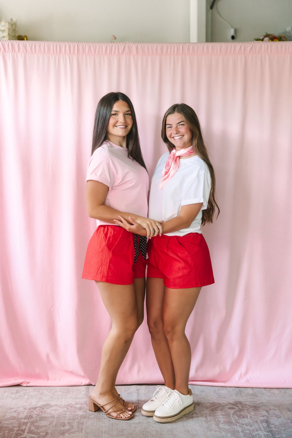 Two women wearing red shorts styled two ways — one with a pink tee and polka dot tie belt, the other with a white tee and red paisley neck scarf