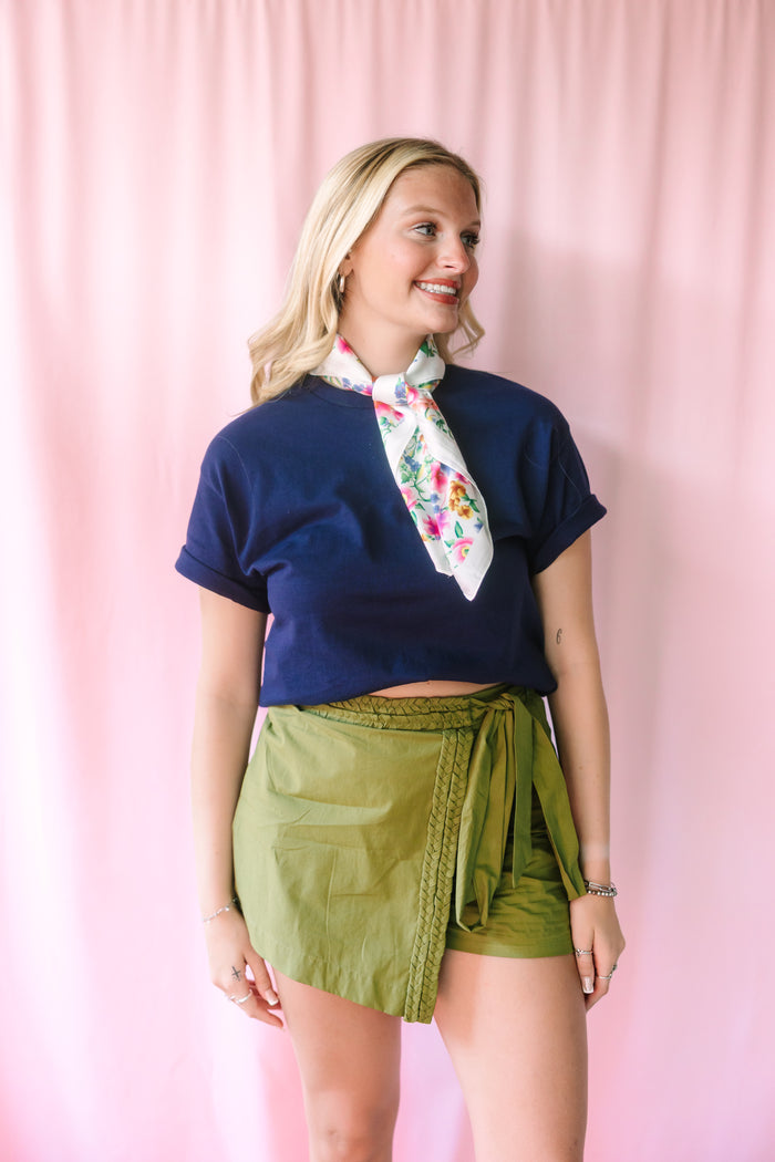 Front view of the Green Braided Trim Skort in olive green featuring a wrap-front design, woven braided trim along the edge, and self-tie bow at the hip, styled with a navy crop tee and floral neck scarf