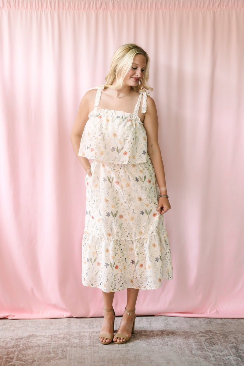 Woman in a cream floral midi dress with tie shoulder straps and tiered ruffle skirt, smiling and looking down against a soft pink curtain backdrop, styled with tan heeled sandals