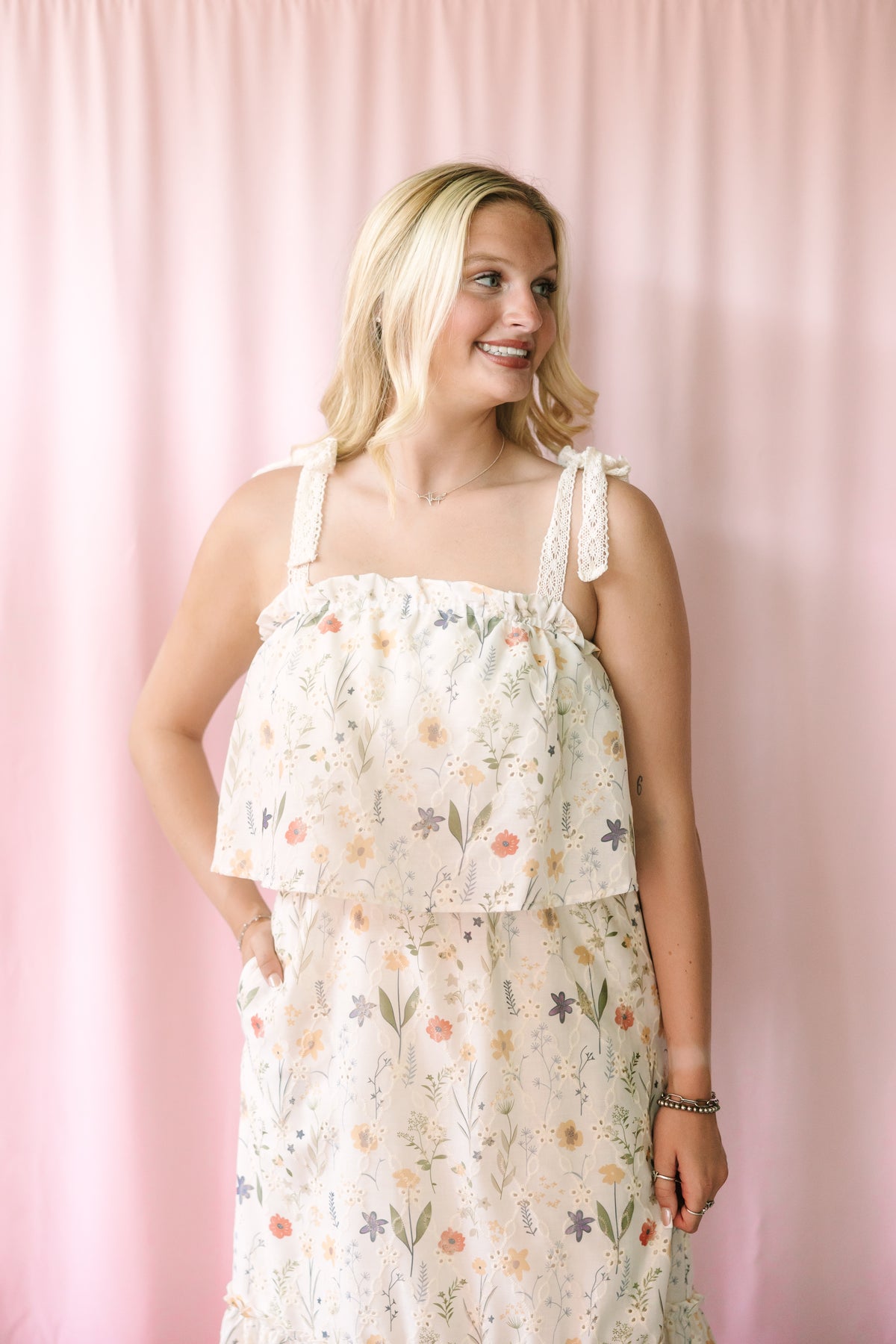 Smiling woman wearing a cream floral sundress with a ruffled overlay bodice and lace tie straps, posed against a blush pink backdrop