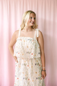 Smiling woman wearing a cream floral sundress with a ruffled overlay bodice and lace tie straps, posed against a blush pink backdrop