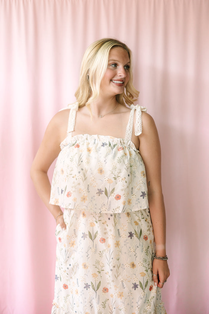 Smiling woman wearing a cream floral sundress with a ruffled overlay bodice and lace tie straps, posed against a blush pink backdrop