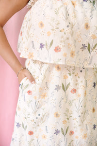 Close-up of a cream floral midi dress showing a tiered ruffle hem and side pocket with a delicate wildflower print in peach, coral, lavender, and sage green
