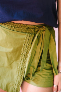 Detail shot of the Green Braided Trim Skort highlighting the woven braided trim along the diagonal wrap-front and elasticized waistband, with fabric self-tie strips at the hip in olive green cotton