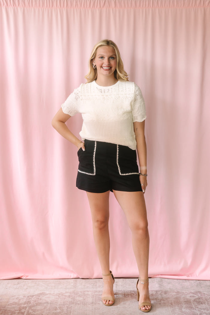 Woman wearing a cream pointelle knit short-sleeve sweater tucked into black high-waist shorts with white zigzag embroidered pocket trim, styled with nude strappy heels against a pink backdrop