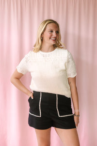 Model smiling in a cream textured knit top and black high-waist shorts with white embroidered pocket detail, photographed against a pink curtain background