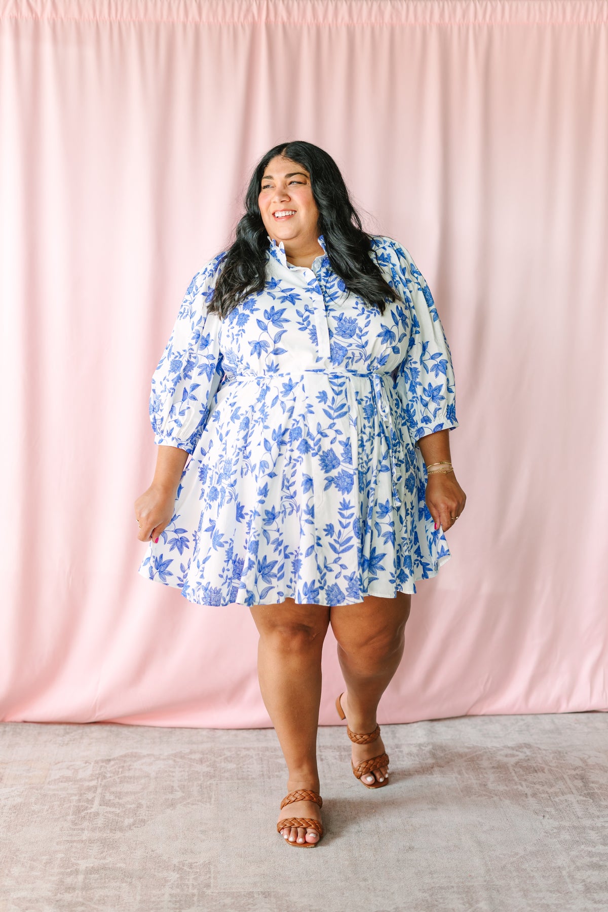 Woman smiling in white mini dress with blue botanical floral print, ruffle neckline, cinched waist tie, and flared tiered hem, styled with tan braided sandals