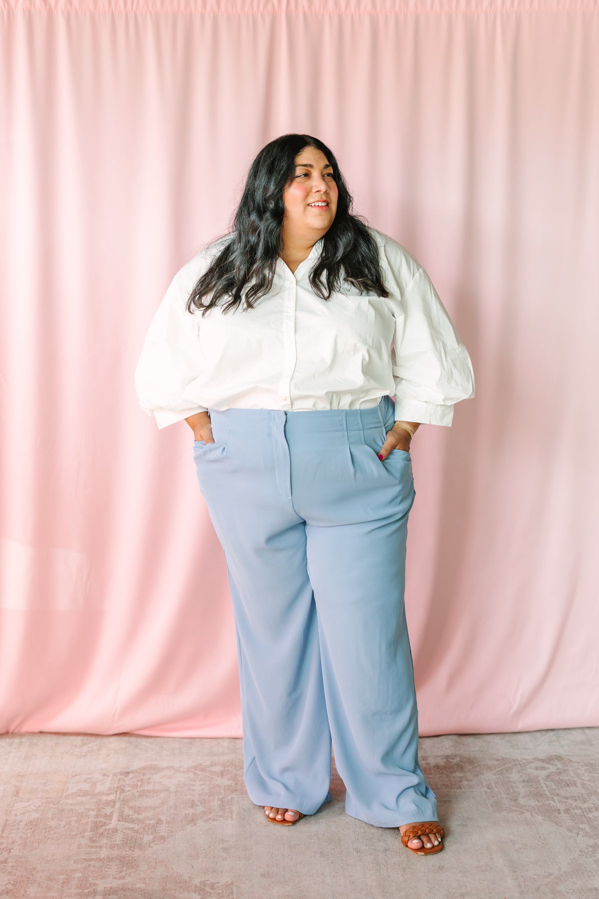 Woman smiling in white poplin button-down top and light blue wide-leg trousers, pink backdrop 