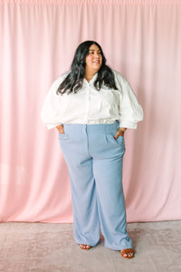 Woman smiling in white poplin button-down top and light blue wide-leg trousers, pink backdrop 