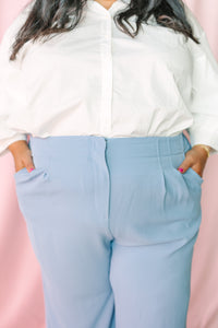 Plus size light blue wide-leg trousers with front pleats and side pockets, paired with a white button-down shirt, pink backdrop
