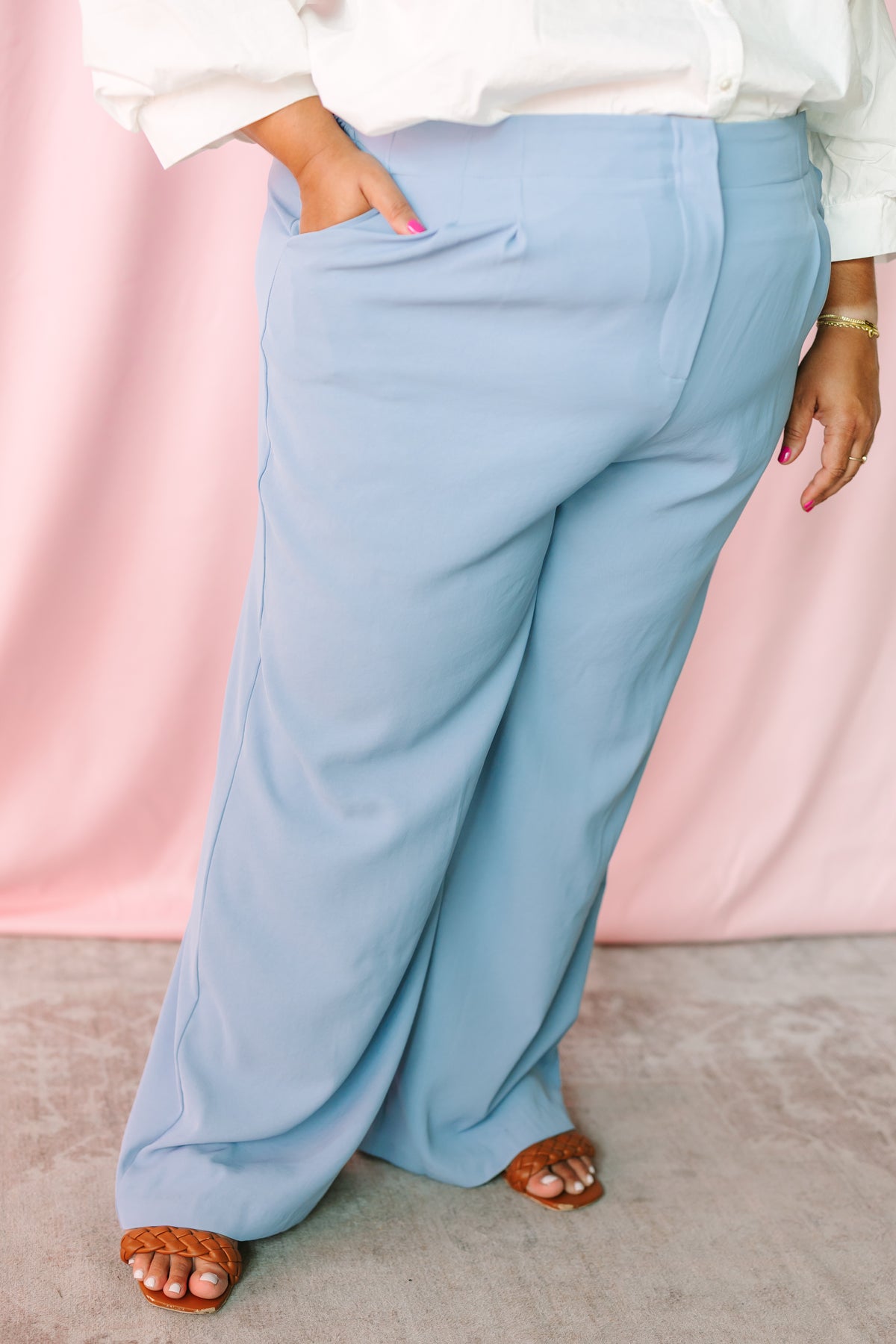 Side view of plus size light blue wide-leg trousers with deep side pocket, paired with a white shirt and tan braided sandals on a pink backdrop