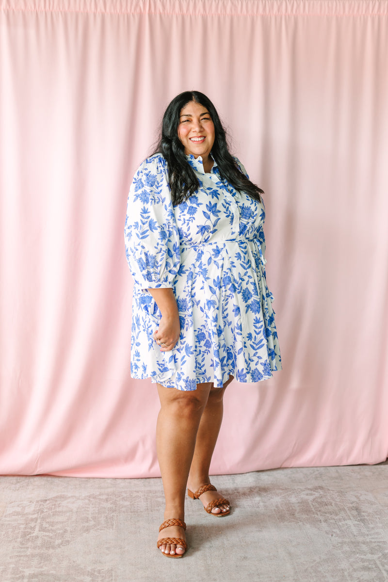 Woman in white blue floral mini dress with 3/4 sleeves, tie waist, and tiered skirt, styled with tan braided block heel sandals