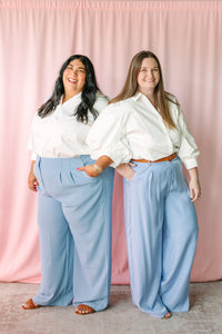 Two women wearing white Chalet Button Down Top styled with light blue wide-leg pants, pink curtain backdrop – Sugarlips inclusive sizing XS–3X
