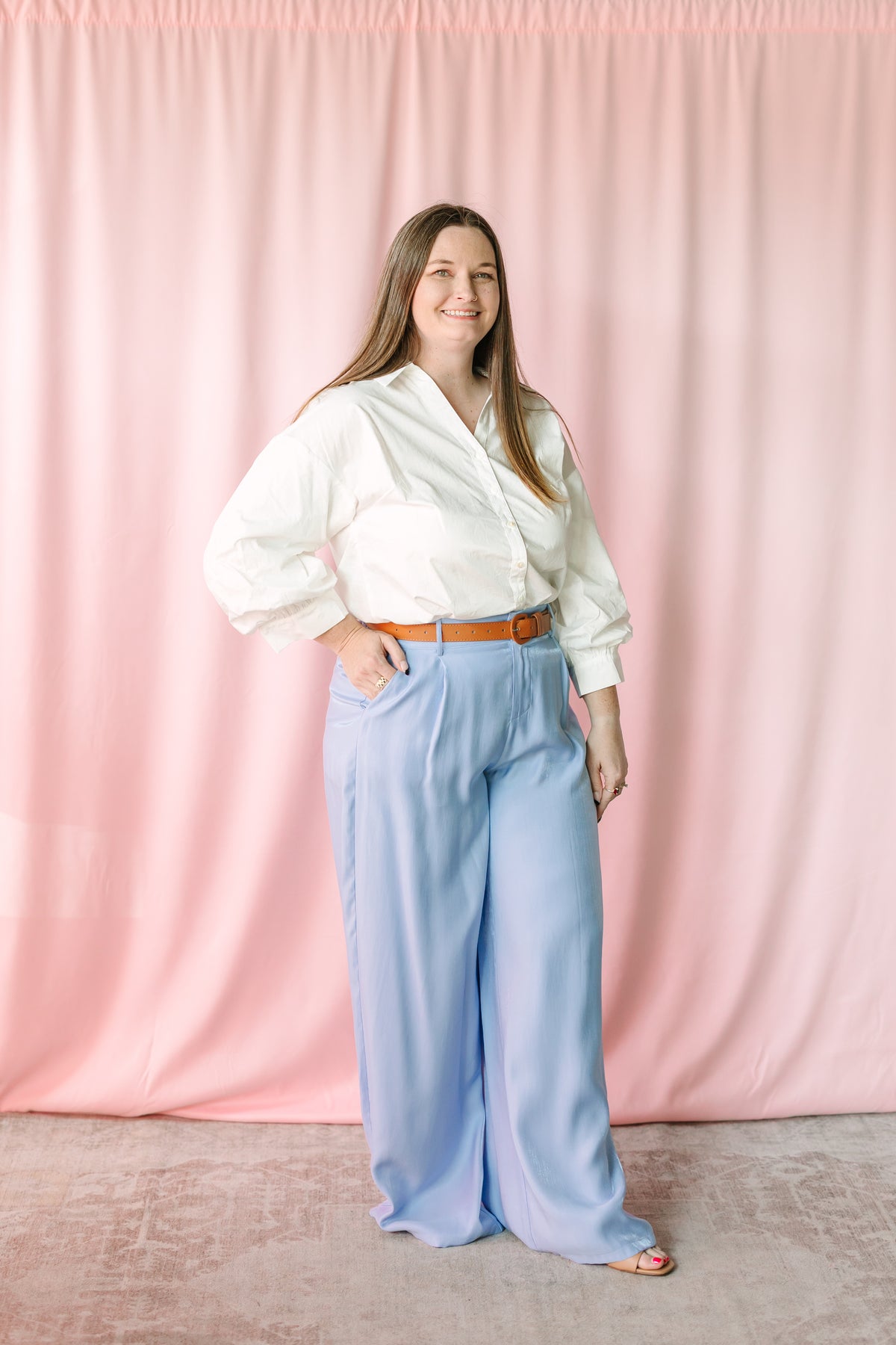 Full-length view of woman in white button-down top and light blue wide-leg trousers, pink backdrop 
