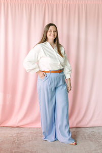 Full-length view of woman in white button-down top and light blue wide-leg trousers, pink backdrop 
