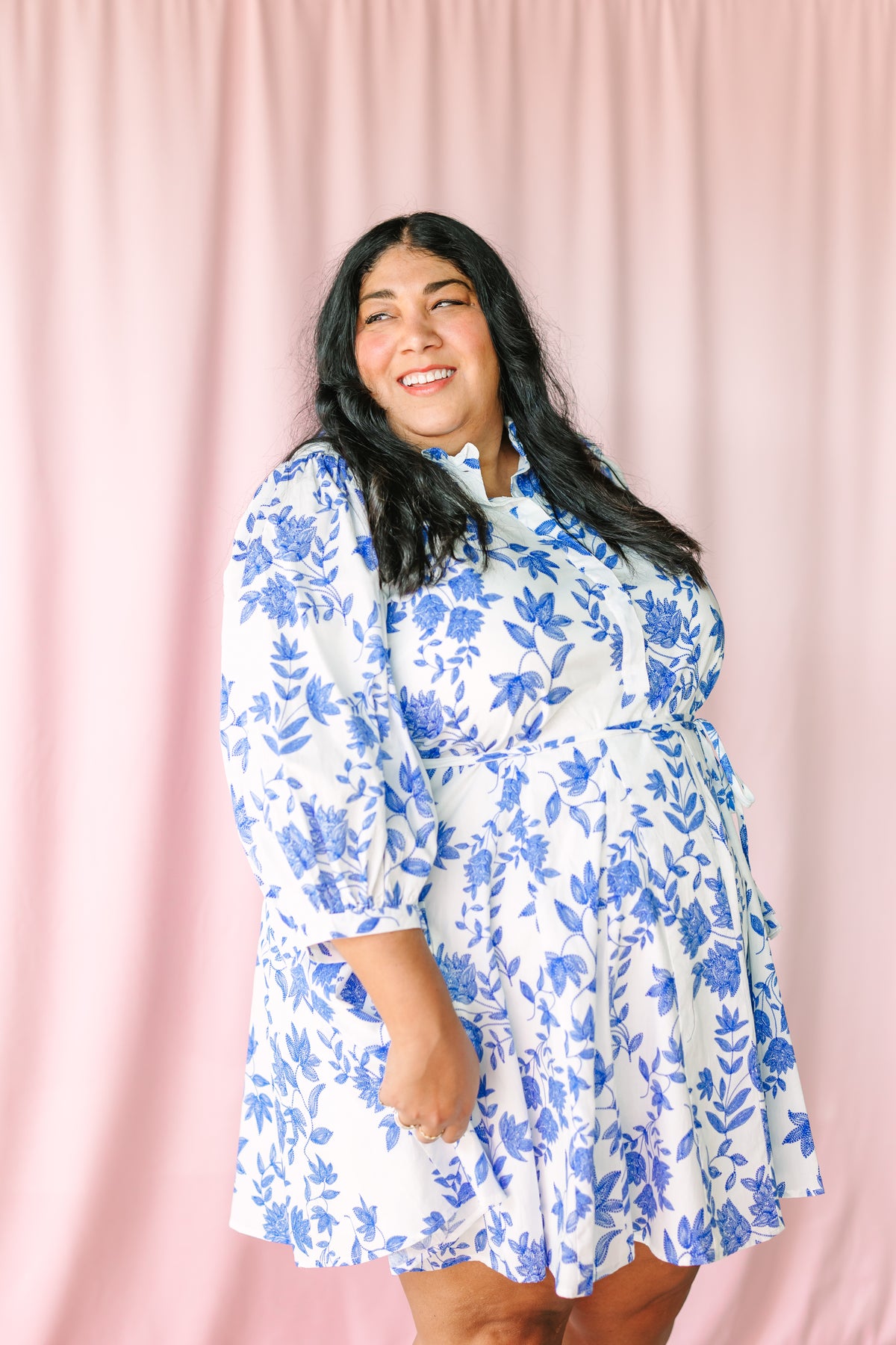 Woman smiling and wearing white mini dress with blue floral print, ruffle collar, tie waist, and flared skirt, against pink curtain backdrop