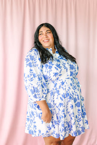 Woman smiling and wearing white mini dress with blue floral print, ruffle collar, tie waist, and flared skirt, against pink curtain backdrop