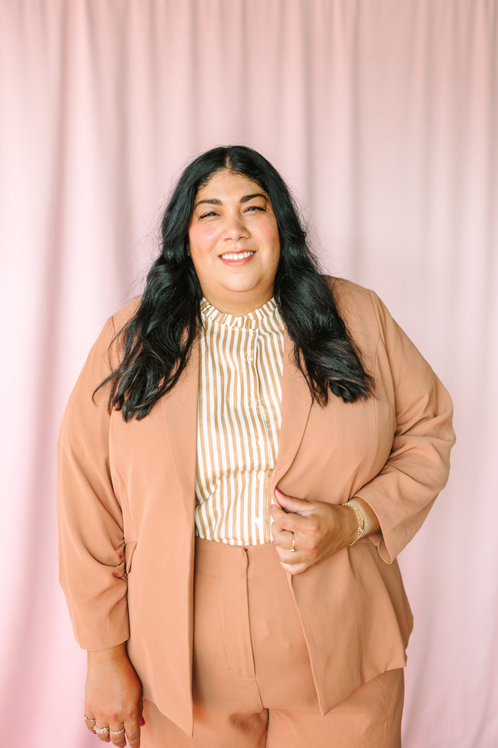 Woman in hazelnut open-front shirred sleeve blazer and gold stripe top smiling, holding blazer lapel, gold bracelet and rings
