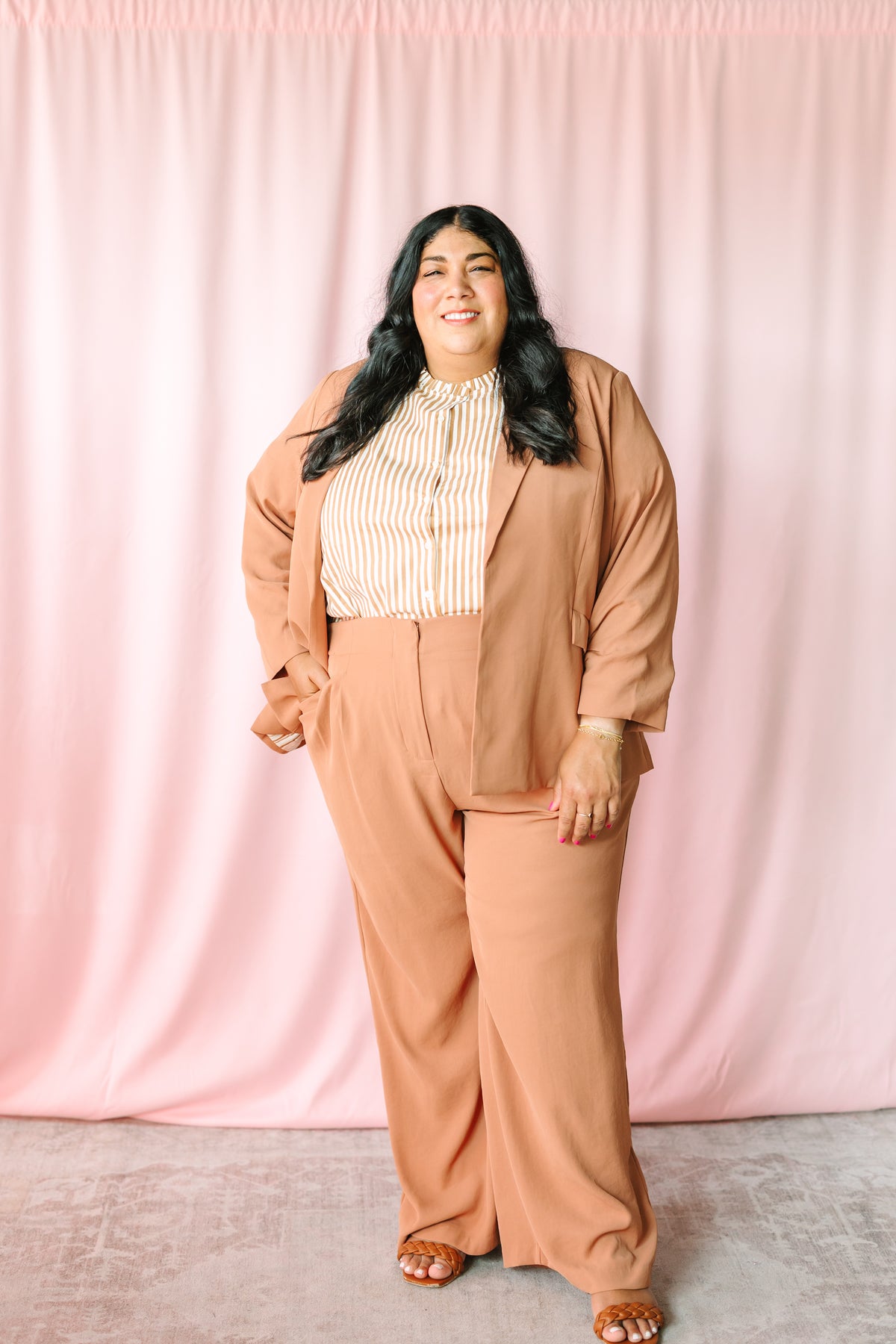 Full-length view of woman in hazelnut shirred sleeve blazer and wide-leg pants with gold stripe top and tan woven heeled sandals on pink background