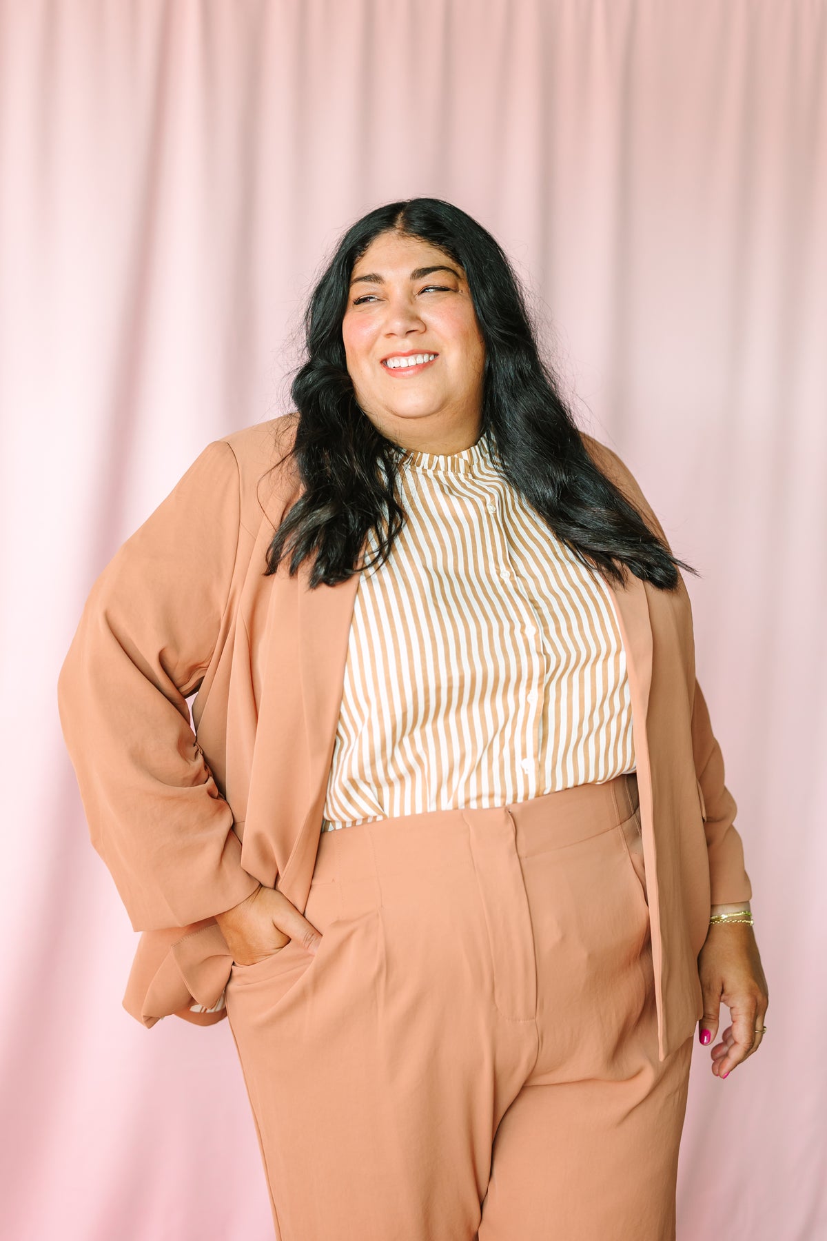 Woman smiling in hazelnut shirred sleeve blazer and wide-leg trousers styled with gold stripe mock-neck top against pink backdrop