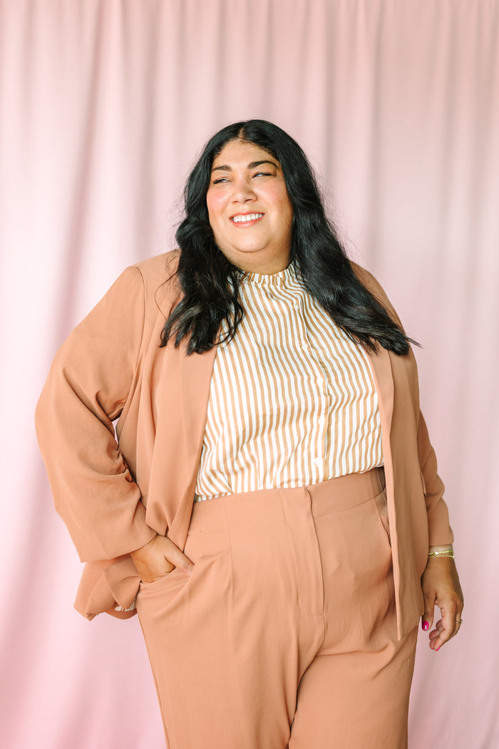 Woman smiling in hazelnut shirred sleeve blazer and wide-leg trousers styled with gold stripe mock-neck top against pink backdrop