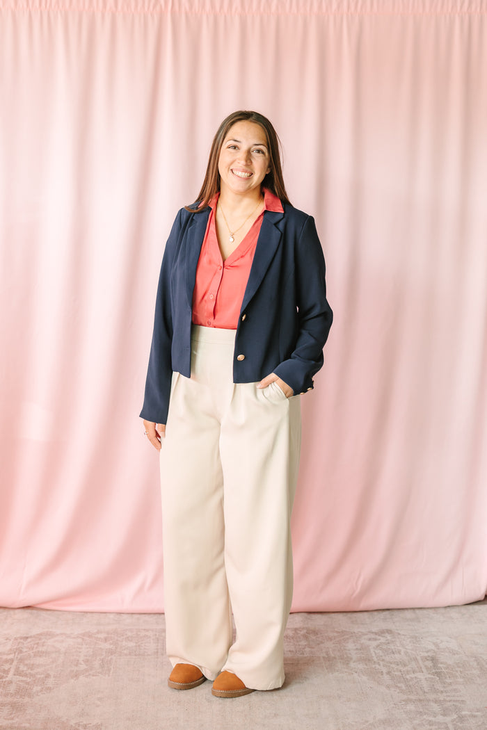 Woman wearing cream wide-leg pleated trousers with a berry blouse and navy cropped blazer with gold buttons, styled with tan platform shoes against a pink draped backdrop