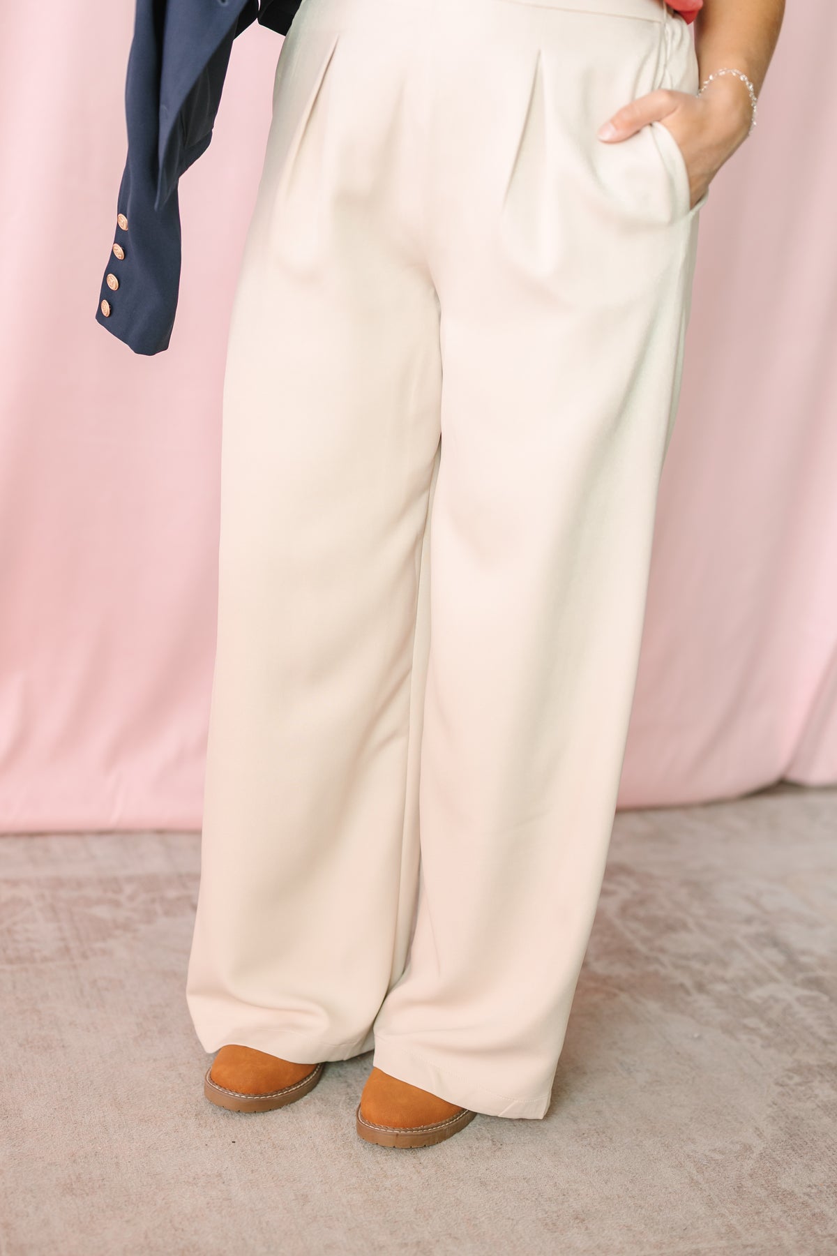 Close-up of cream wide-leg pleated trousers with side pockets, styled with tan platform shoes against a pink draped backdrop