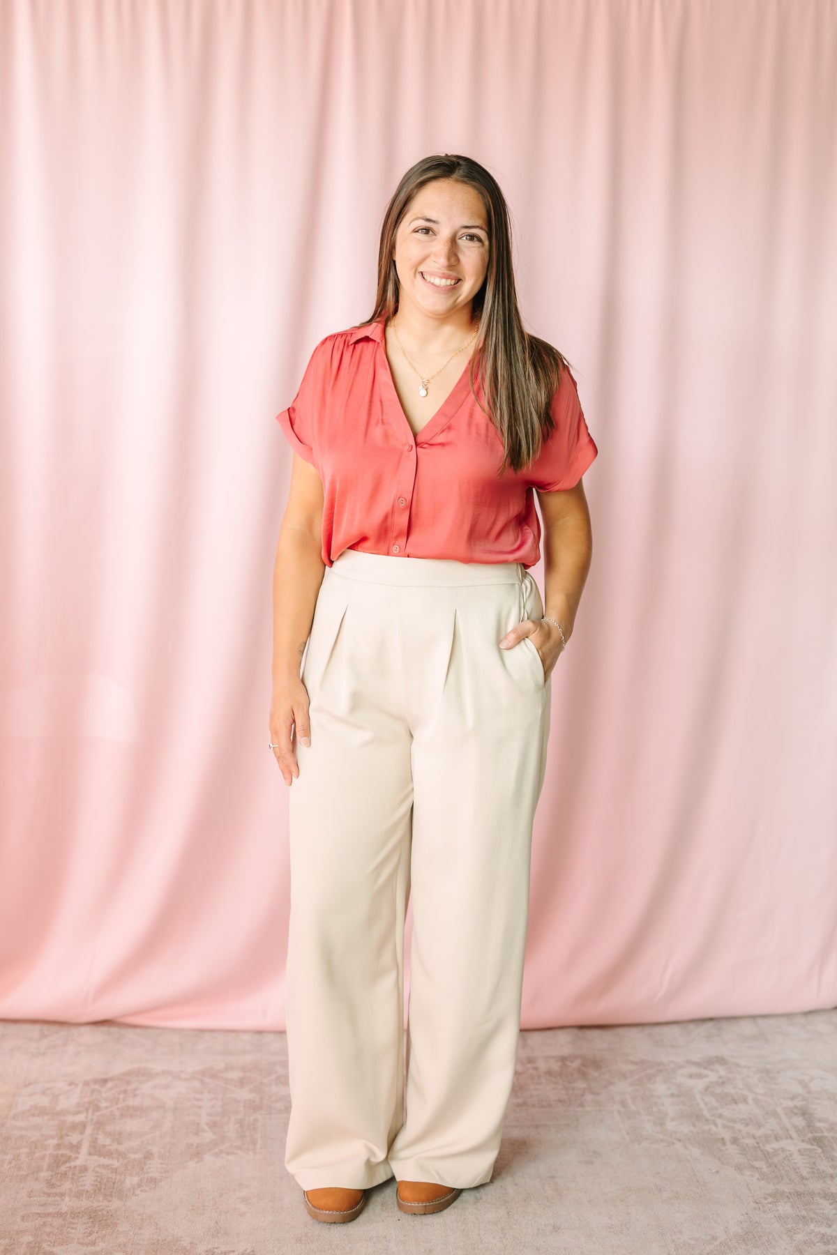 Woman wearing cream wide-leg pleated trousers with a berry short-sleeve button-down blouse, styled with tan platform shoes against a pink draped backdrop