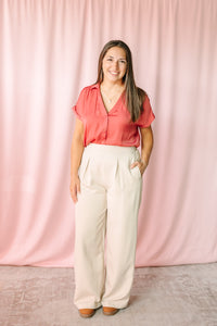 Woman wearing cream wide-leg pleated trousers with a berry short-sleeve button-down blouse, styled with tan platform shoes against a pink draped backdrop