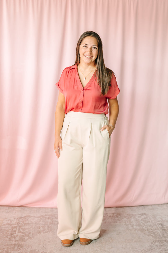 Woman wearing cream wide-leg pleated trousers with a berry short-sleeve button-down blouse, styled with tan platform shoes against a pink draped backdrop