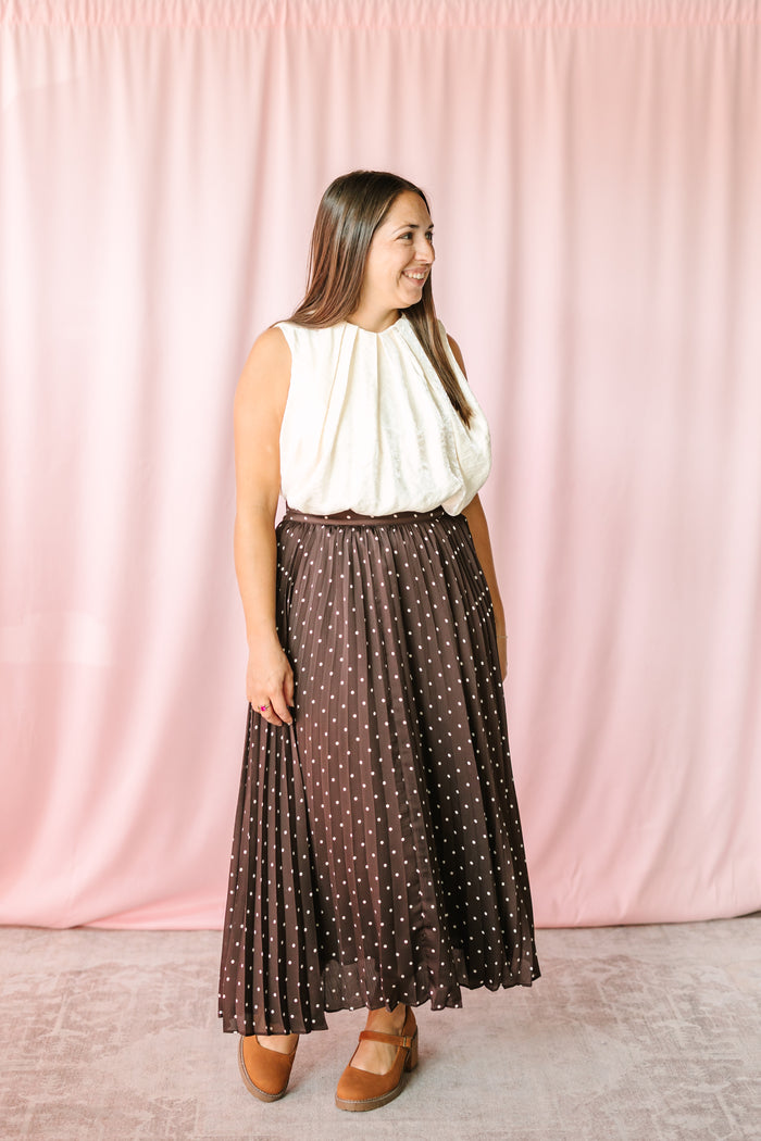 Woman wearing chocolate brown polka dot pleated maxi skirt paired with cream sleeveless gathered top and tan Mary Jane wedges against pink backdrop