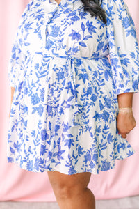 Close-up of white mini dress with vibrant blue botanical floral print and tie waist detail, showing flared tiered skirt