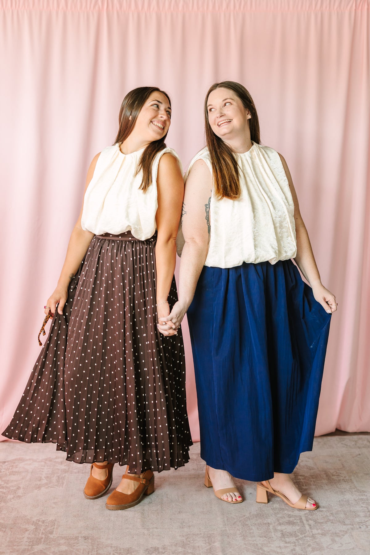 Two models wearing cream sleeveless pleated balloon top — styled with navy wide-leg skirt and brown polka dot midi skirt