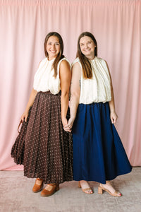 Two women wearing pleated maxi skirts — chocolate brown polka dot and solid navy — both styled with cream sleeveless tops against pink backdrop