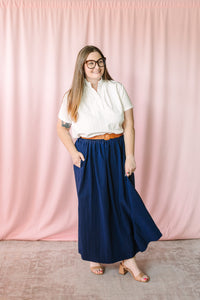 Woman wearing a white short-sleeve button-front top with a mandarin collar, navy blue maxi skirt with pockets, tan leather belt, and nude block-heel sandals, standing against a soft pink draped backdrop