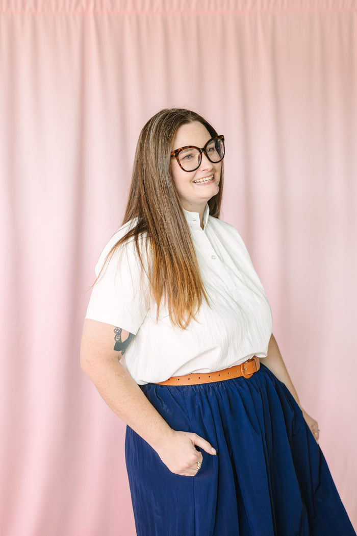 Woman in tortoiseshell glasses smiling in a three-quarter pose, wearing a white short-sleeve button-front top, navy blue gathered maxi skirt with pockets, and tan leather belt against a pink draped background