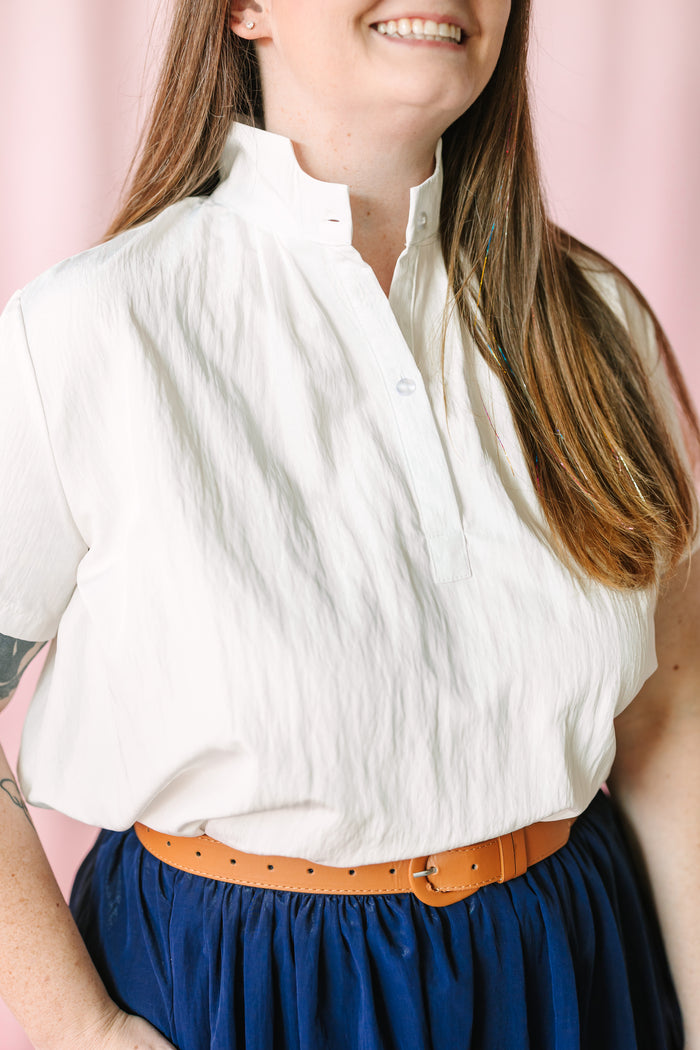Close-up of a white short-sleeve linen-blend button-front top with mandarin collar, tucked into a navy blue gathered maxi skirt with a tan leather perforated belt