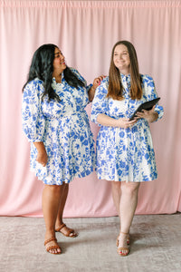 Two women smiling and wearing matching white mini dresses with blue floral print, 3/4 sleeves, and tie waist, styled with tan heeled sandals against a pink backdrop